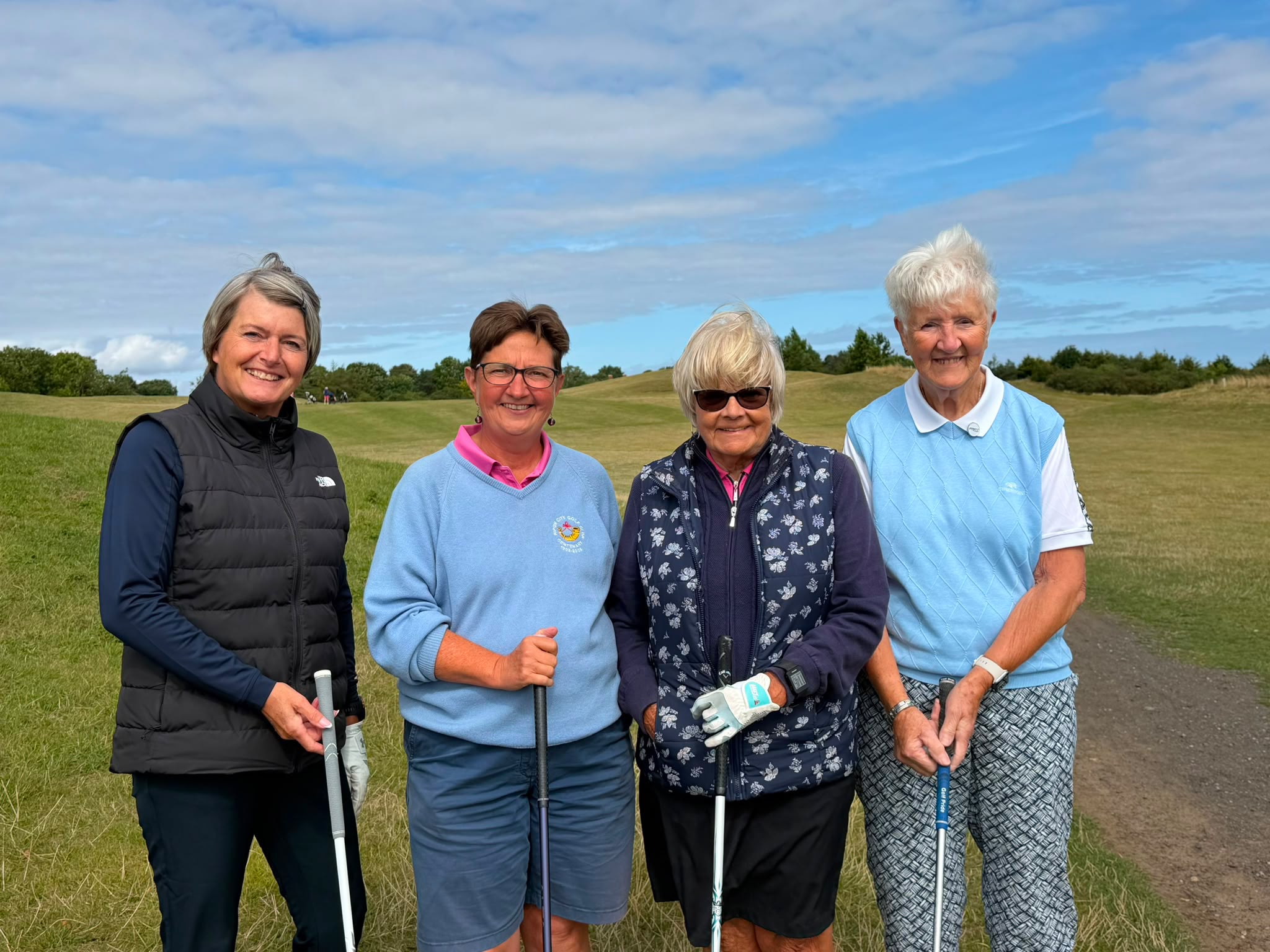 Four Cleveland Golf Club lady members smiling together beside the fairway, with rolling greens and a clear sky behind them
